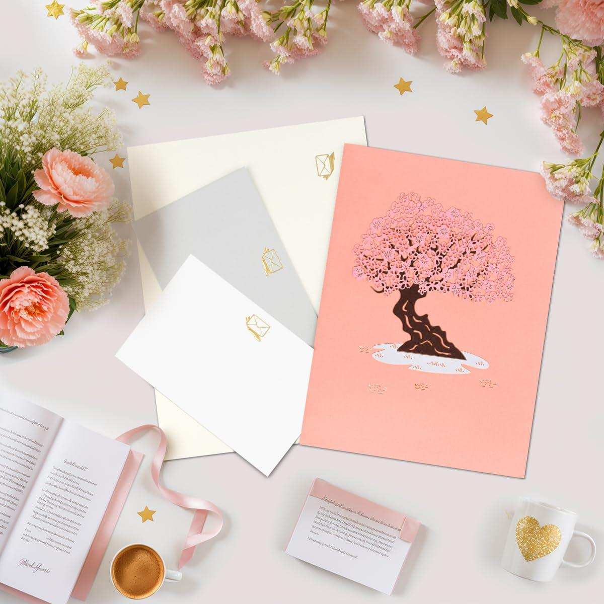 Pink pop-up greeting card with cherry blossom tree, envelopes, flowers, and mug on desk TUTT