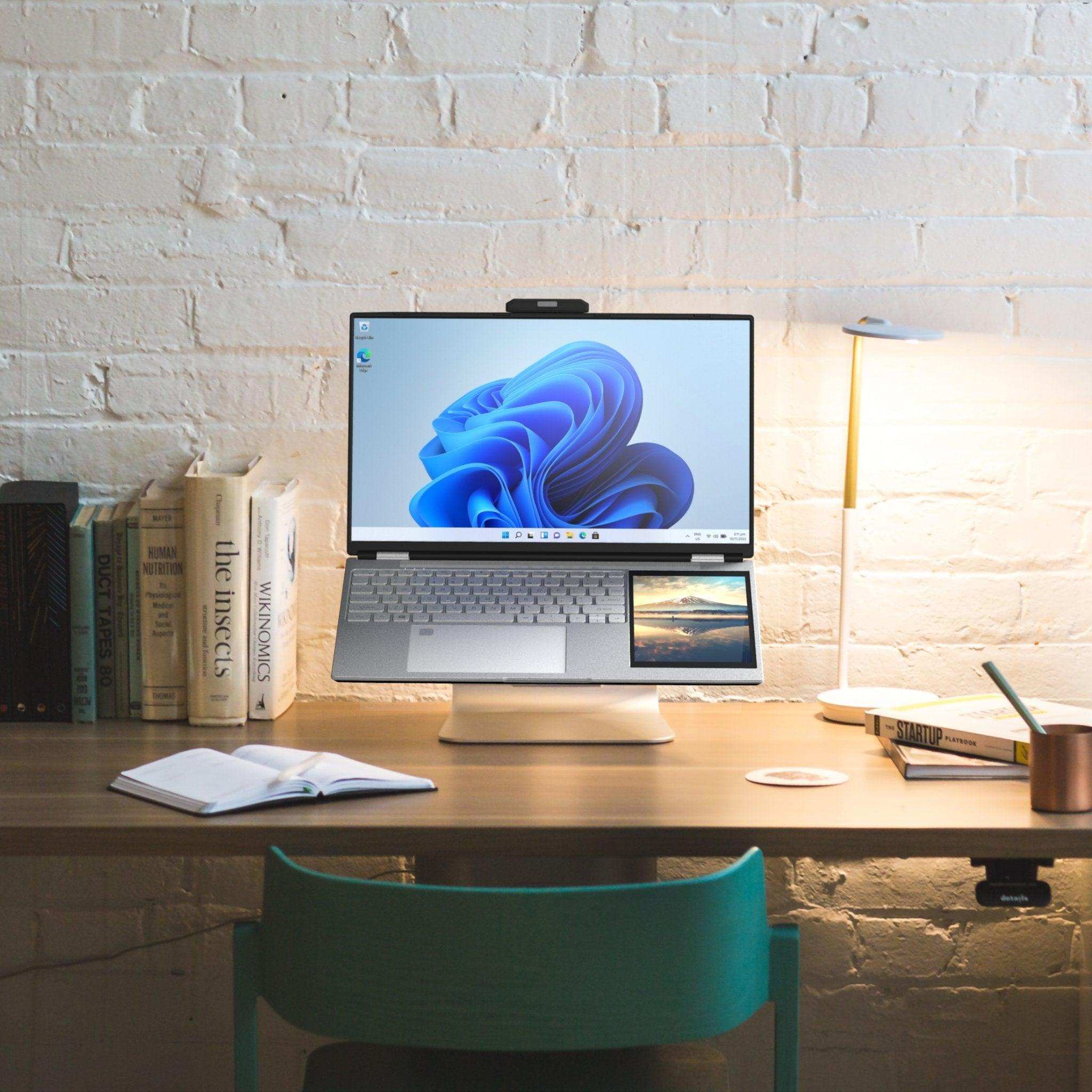 Silver dual-screen laptop with mini touchscreen on a modern desk setup, books, and lamp. TUTT