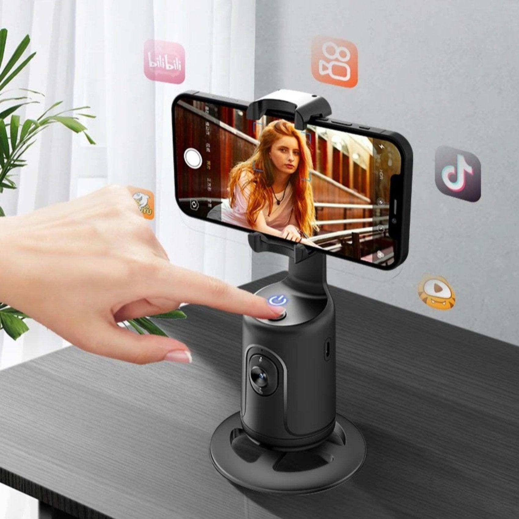 Smartphone mounted on black 360° auto face tracking selfie holder gimbal on desk, hand pressing button, social media icons visible, TUTT